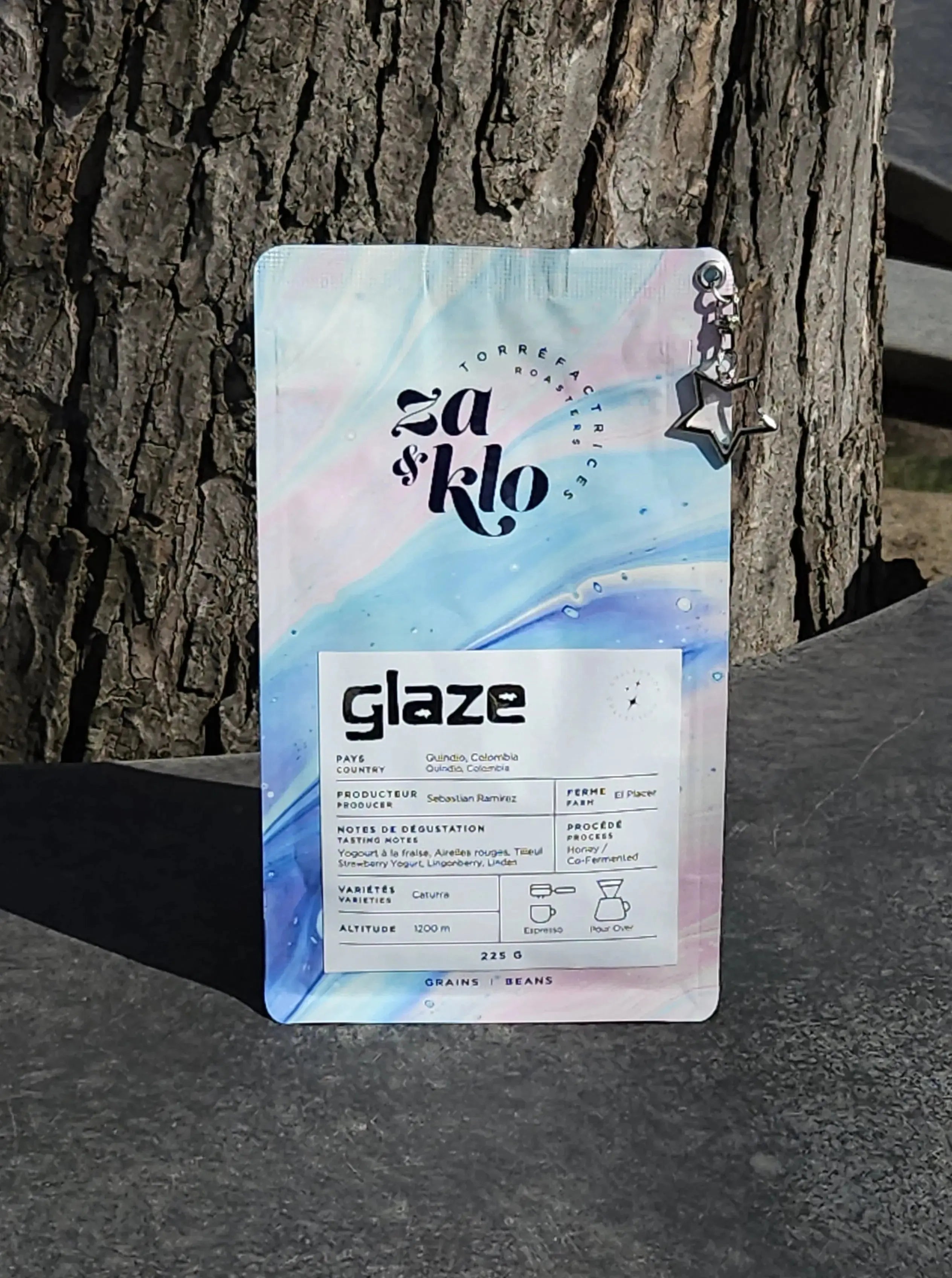 Photo of Glaze, a 'honey' co-fermented coffee from Colombia produced by Sebastian Ramirez, with a star-shaped keychain attached to the bag. The bag highlights fruity notes of strawberry yogurt, cranberries, and linden, with visible typography displaying the name 'Glaze.' The 120-hour anaerobic fermentation with red wine yeast, dehydrated strawberries, and red fruits create a texture reminiscent of autumn honey. Ideal for espresso or pour-over. Roasted by Za and Klo Roastery