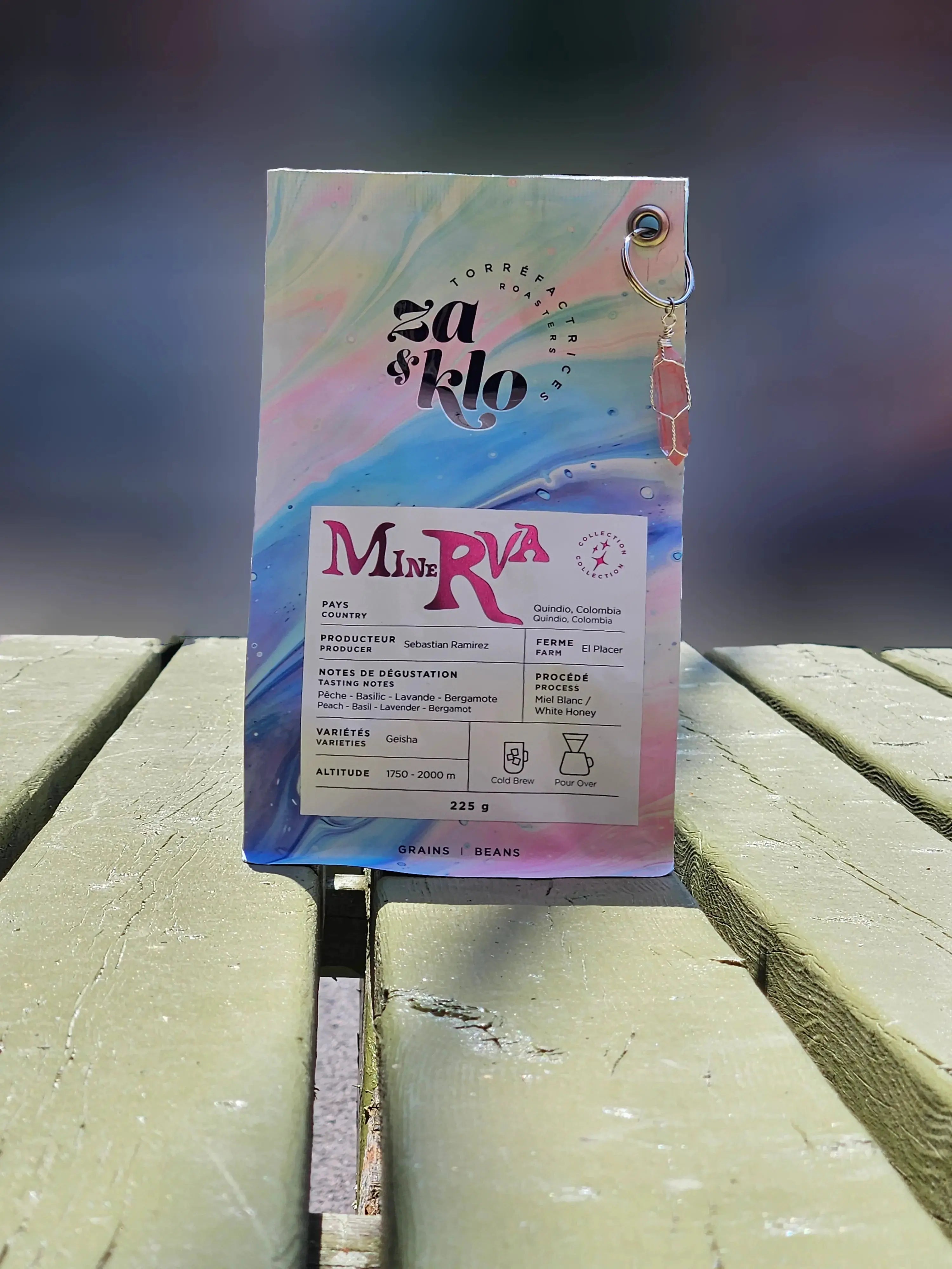 Photo de Minerva, un cafe Geisha « white honey » de Colombie produit par Sebastian Ramirez, avec un porte-clés en cristal attaché au sac. Le sac met en avant des notes fruitées, florales et herbacées, avec une typographie visible affichant le nom « Minerva ». Ce café complexe et surprenant révèle des saveurs de pêche et de basilic, idéal pour accompagner une burrata ou une pâtisserie aux fruits. Parfait en cold brew ou pour over. Torrefactrices Za et Klo.